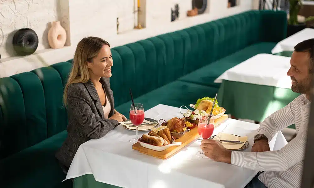 A long run of velvet green deep fluted banquette seating in a cafe, a couple is sat having a meal