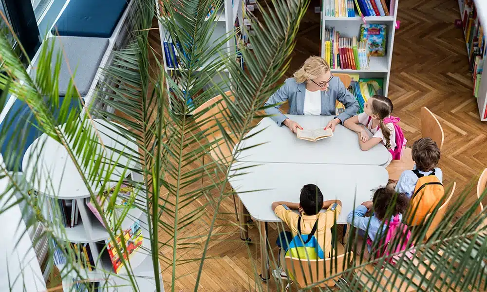 a lady sits in a library with school children around a large white laminate table top