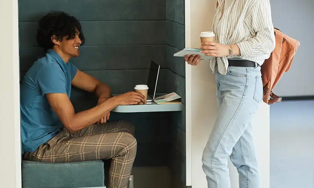 Single seater solo office pod with a man working infront of a table, a woman stands alongside with documents and a coffee