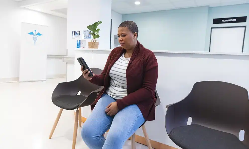 A patient at a UK gp clinic sits on an upholstered plastic chair in a waiting room