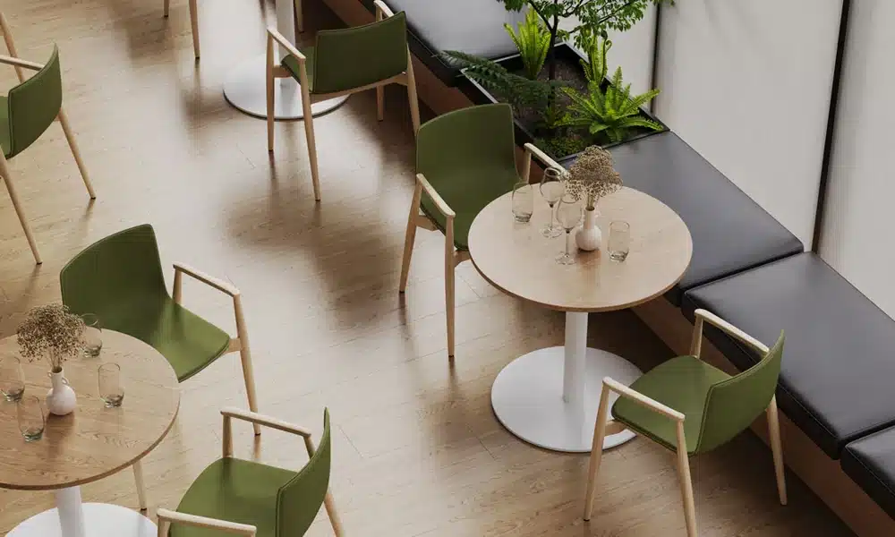 Scandinavian style restaurant cafe with light oak table tops and white bases on the floor, green upholstered chairs and grey bench seating in the background