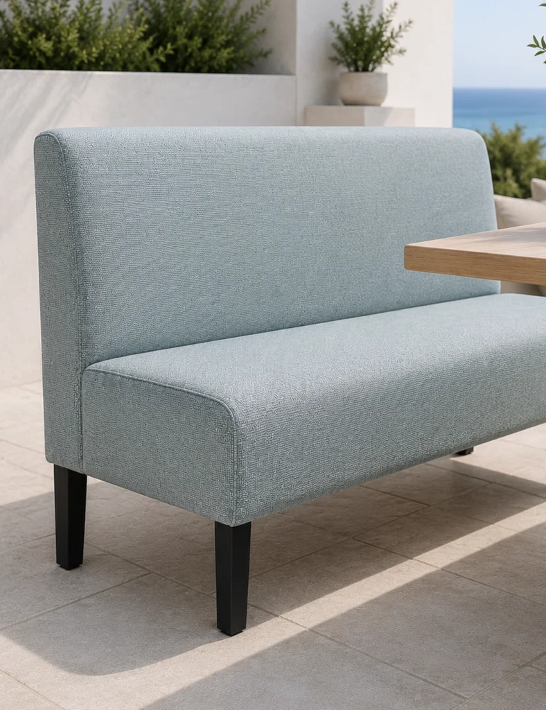 Outdoor seating: light blue upholstered bench with black legs on tiled patio, wooden table beside, ocean in background