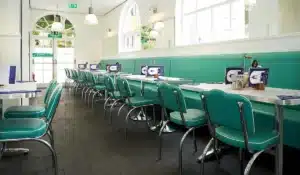 Retro diner interior with teal vinyl booths and chairs along a long white table, large arched windows, and pendant lighting.